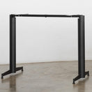 Switch The Parkway Black Self Levelling Table Base - Twin Pedestal - NOROCK features a black metal frame with two sturdy legs and a horizontal support bar, positioned on a polished concrete floor against a plain white wall, without a tabletop. 2 image