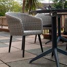 Switch The Hanover Outdoor Stacking Tub Chair with black legs is placed next to a round outdoor table on a tiled patio, set against a wooden fence with trees and houses in the background. 2 image