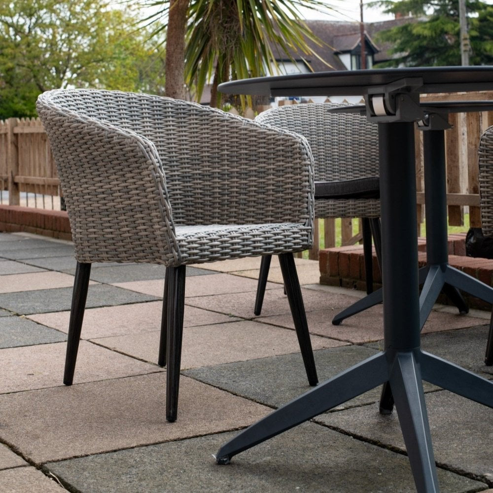 The Hanover Outdoor Stacking Tub Chair with black legs is placed next to a round outdoor table on a tiled patio, set against a wooden fence with trees and houses in the background.