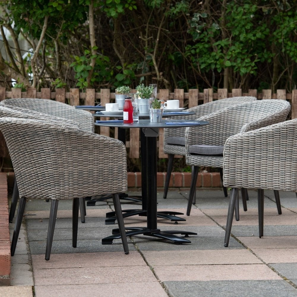 Four Hanover Outdoor Stacking Tub Chairs surround a round outdoor table with coffee cups, plates, and ketchup. The paved area is bordered by green bushes and a wooden fence.