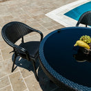 Switch A Paris Bistro Outdoor Stacking Armchair and a round glass-topped table sit on a stone patio by the pool, with a fruit basket containing bananas and grapes on the table. 3 image