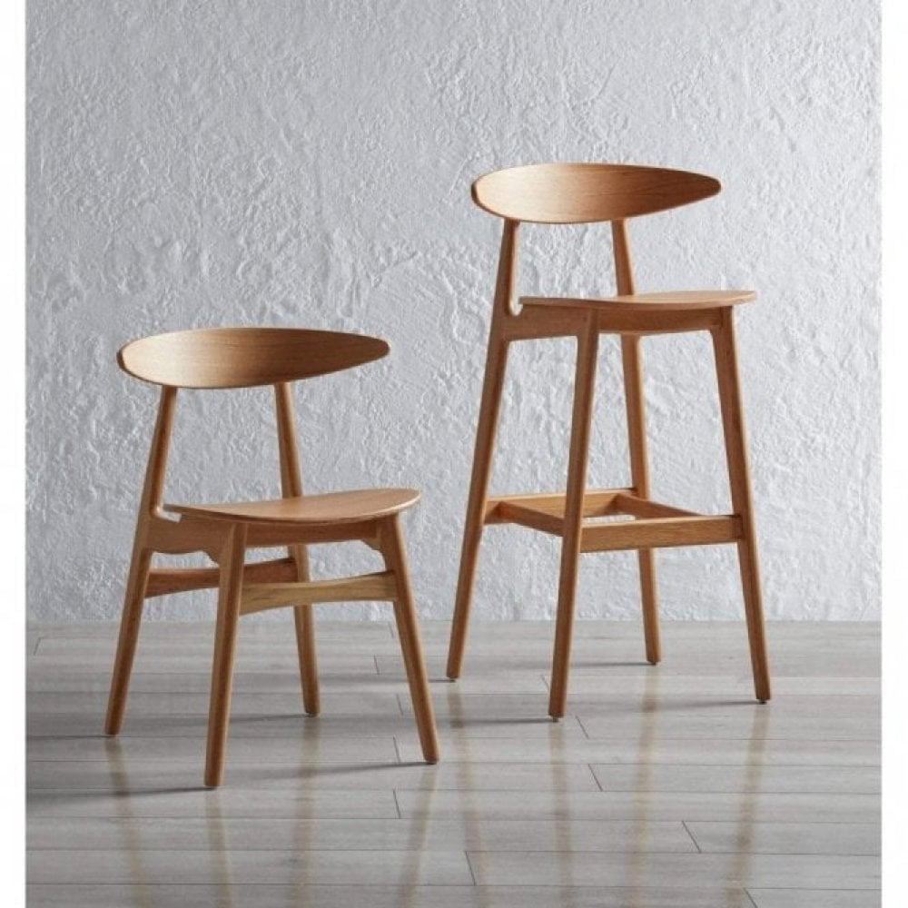 The Carcher Oak Bar Stool, with its minimalist design and curved backrest, crafted from solid oak, stands on a light wooden floor and enhances the contemporary interior against a textured white wall.