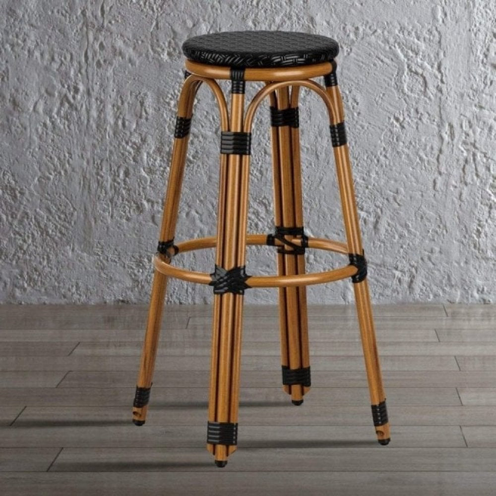 The Time Bistro Outdoor Stacking Bar Stool features a tall black wicker frame, round woven seat, and black wrapped leg accents, standing on a light brown wooden floor against a textured gray wall.