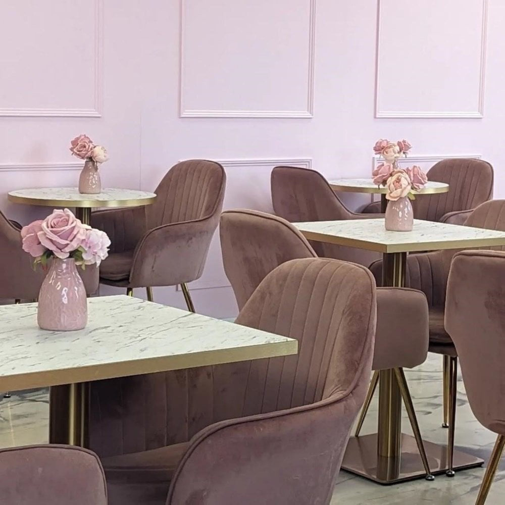 A chic cafe featuring pink velvet chairs and Zeus Brass Base Pedestal Square Tables with white marble effect tops (700x700mm), each adorned with a small vase of pink roses, set against a pastel pink wall.