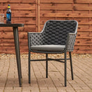Switch The Cordoba Outdoor Stacking Armchair, with a textured dark gray woven backrest and seat cushion on an aluminum frame, sits beside a dark round table holding a bottle and glass on a stone patio in front of a wooden fence. 3 image