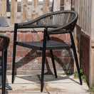 Switch The Vienna Outdoor Stacking Rattan Armchair features a woven rope backrest and seat, black metal legs, and a curved design, shown on a stone patio near a wooden fence and brick wall. 2 image