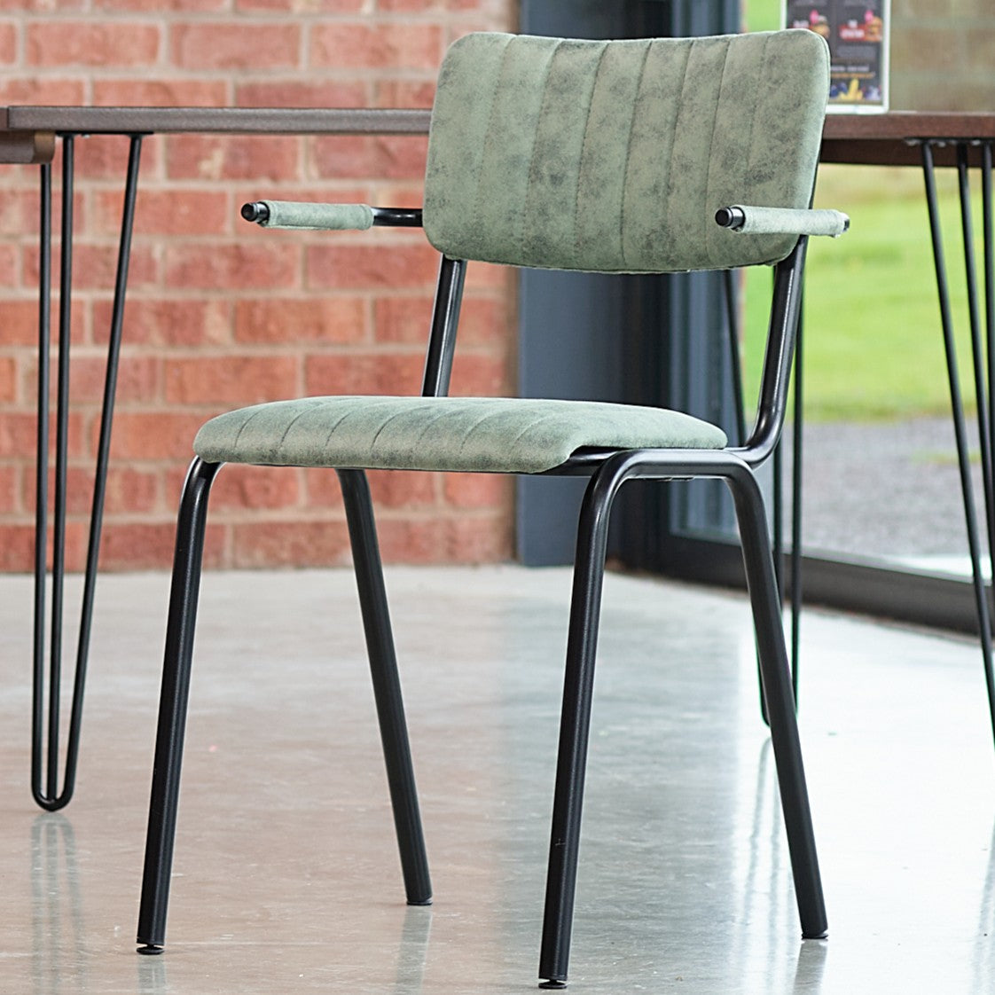 The Derby Industrial Armchair features a light green faux suede padded seat and backrest, black metal frame with armrests, set on a polished concrete floor beside a brick wall and large windows.