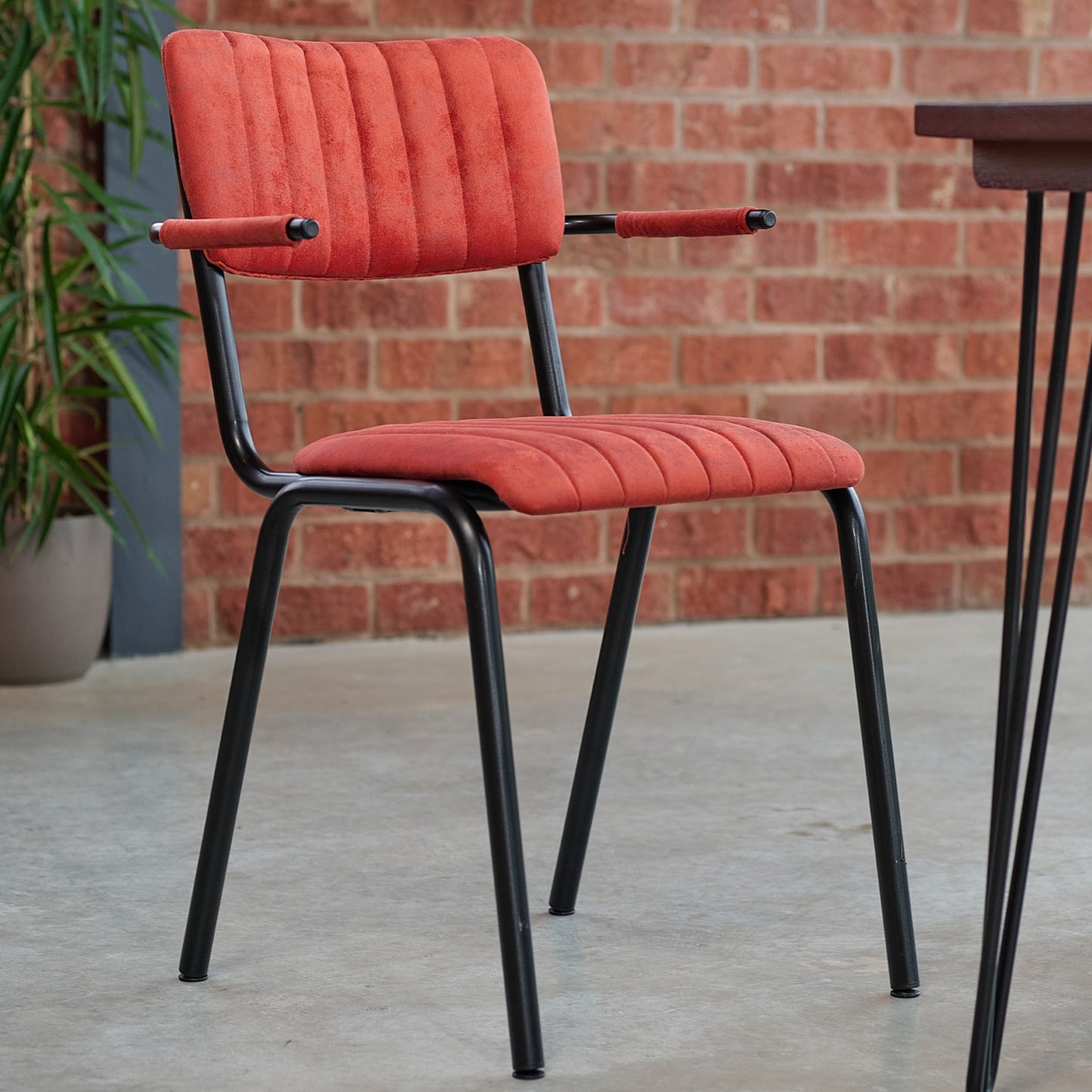 The Derby Industrial Armchair features a red cushioned faux suede seat and backrest, black metal frame with armrests, set on a concrete floor beside a brown brick wall and a potted plant.