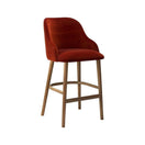 Switch The Onyx Grande Barstool Frame features a modern design with a curved, upholstered red velvet seat and backrest, set on four light solid wood legs with a wooden footrest, displayed against a white background. 2 image