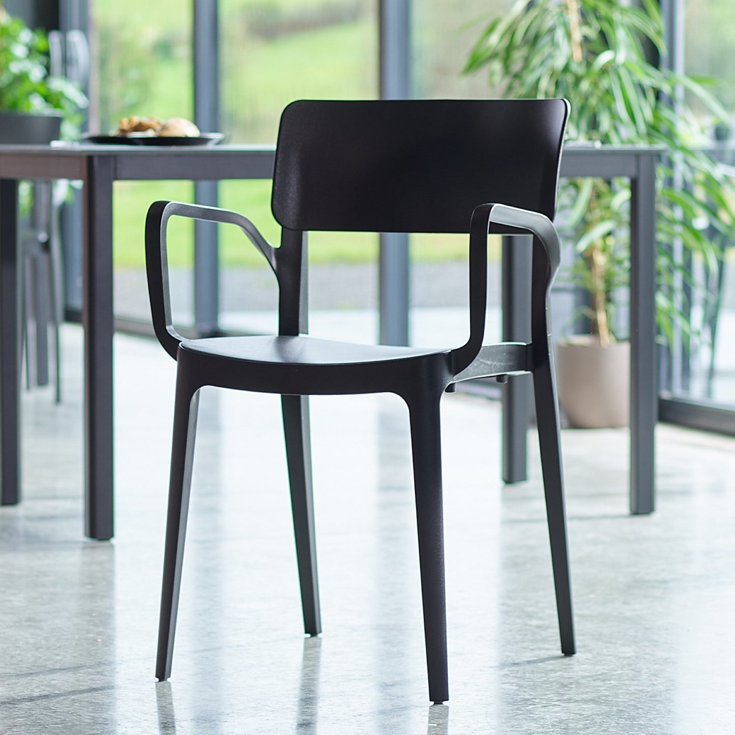 The Vivo Stacking Outdoor Armchair in modern black, featuring armrests, sits on a polished concrete floor in a bright room with large windows, a table, and green plants—a perfect blend of indoor-outdoor chair style.