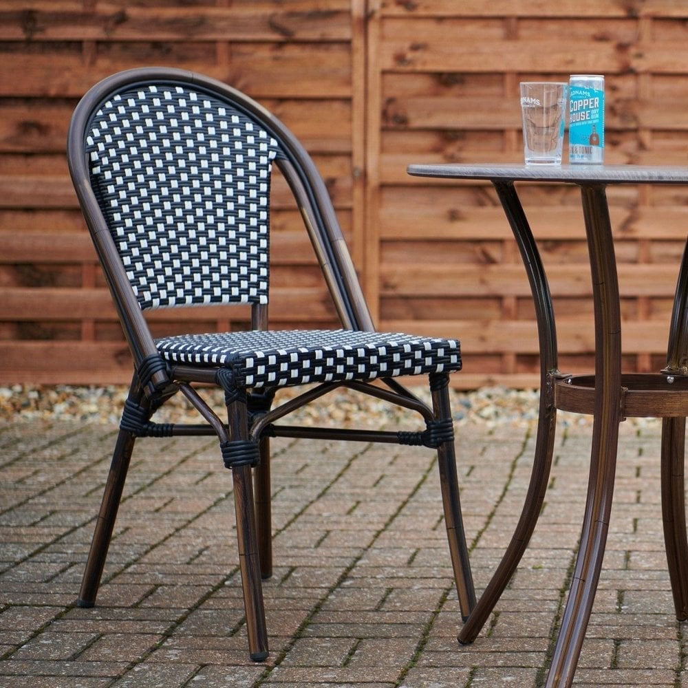 The Carcassonne Bistro Outdoor Stacking Side Chair, in woven black and white with an aluminum frame, sits by a round glass table holding a glass, book, and drink can on a paved patio in front of a wooden fence.