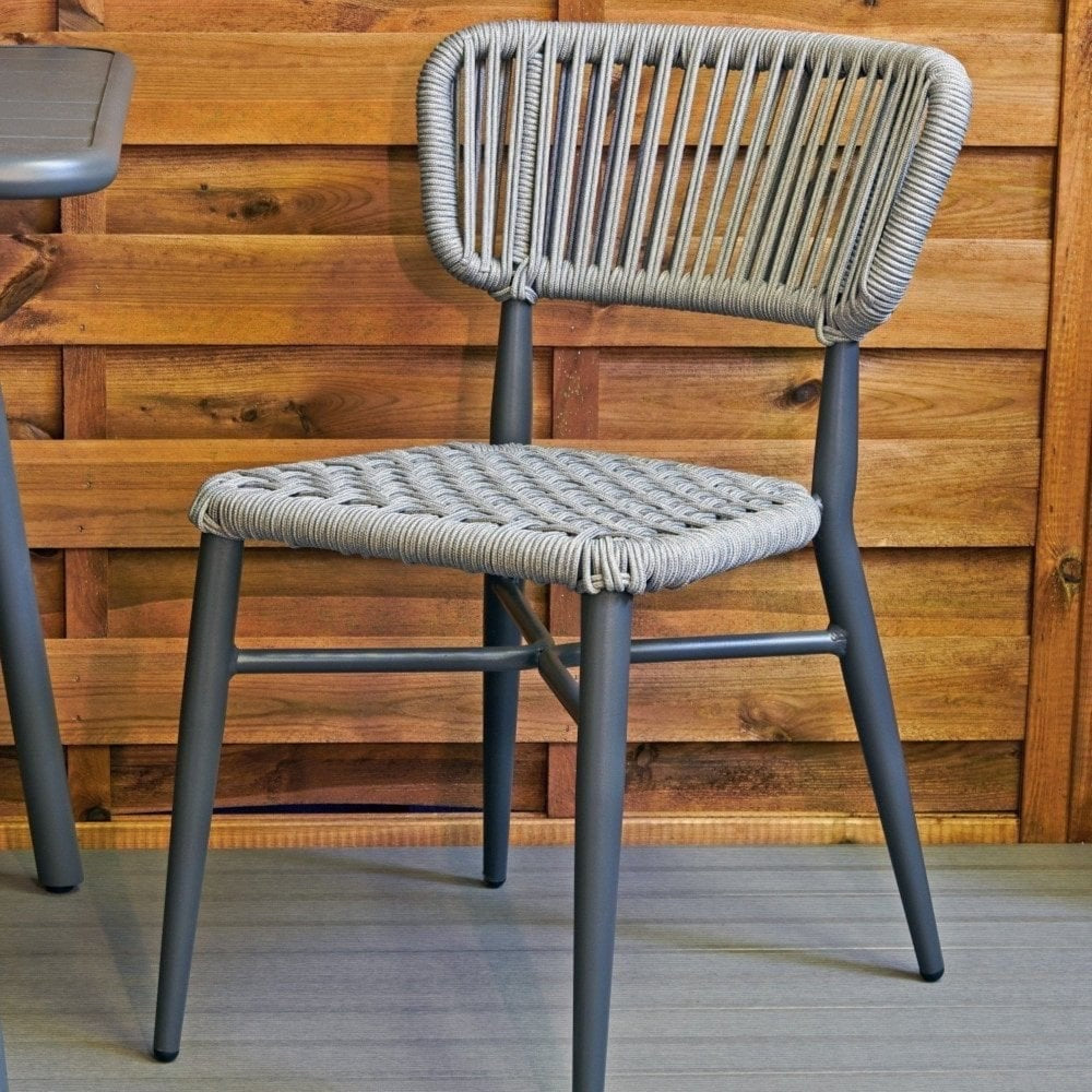 The Madrid Outdoor Stacking Rattan Chair, featuring a woven polyester rope seat and backrest with metal legs in gray, is placed on a patio with a wooden fence in the background.