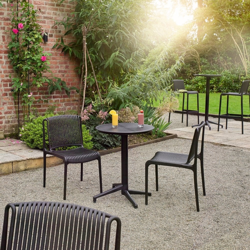 The Boom Flip Bistro Round Table 600mm Round is set with two black chairs on a gravel patio, featuring yellow and red bottles, surrounded by green plants and a brick wall, with sunlight filtering through the trees.