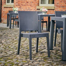 Switch The Canterbury Outdoor Rattan Stacking Side Chair is set on a cobblestone patio outside a brick building with green-framed windows. Flower arrangements on tables create inviting, low-maintenance garden seating. 2 image